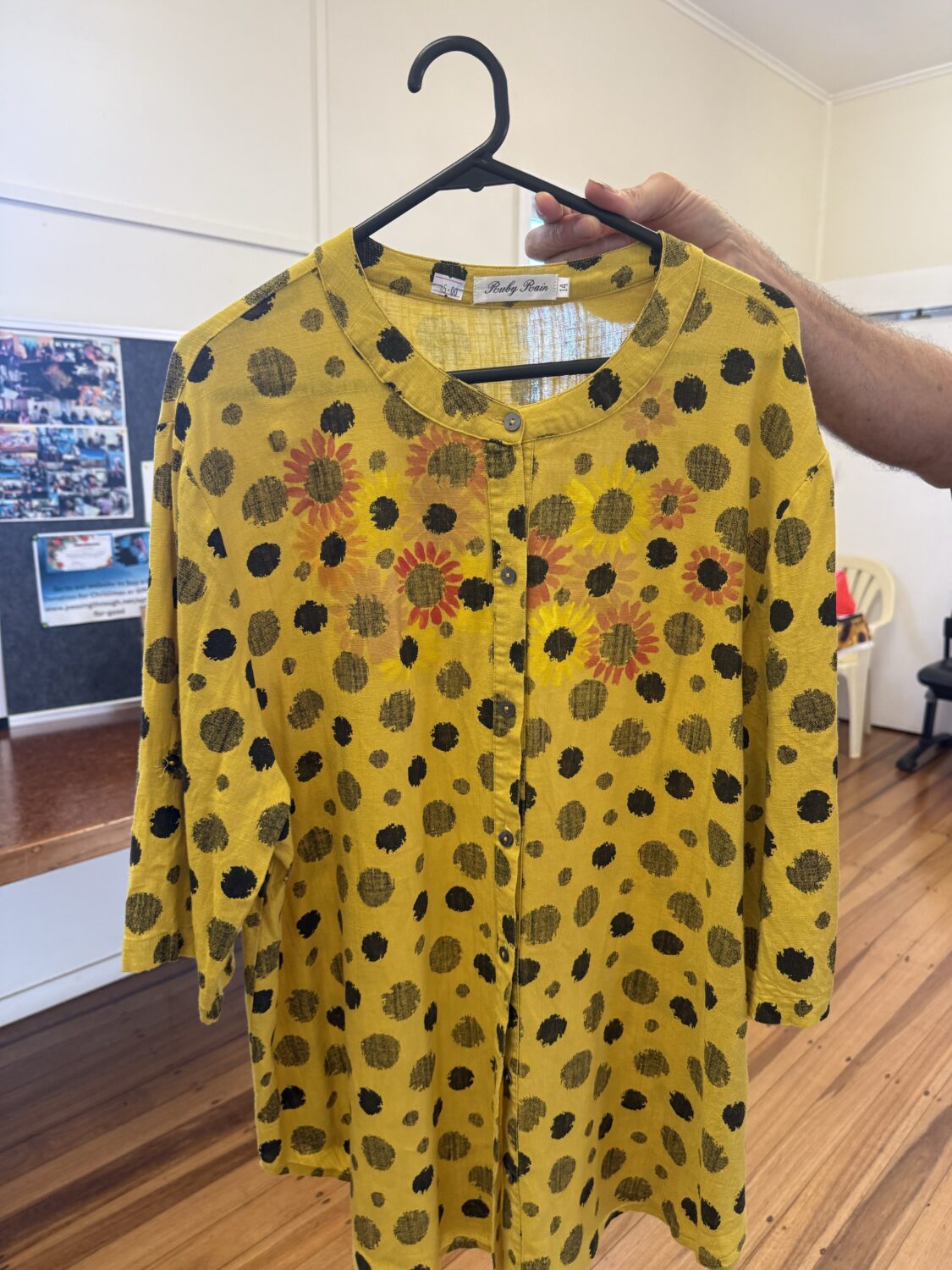 Yellow dress with black polka dots and orange sunflowers on a black hanger, held by a hand, button-front detail visible.