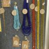 Beaded necklace display with turquoise, royal blue, orange, and yellow strands; sunflower charms and price tags on a gray board.