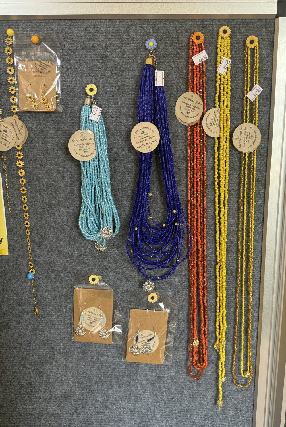Beaded necklace display with turquoise, royal blue, orange, and yellow strands; sunflower charms and price tags on a gray board.