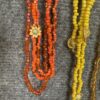Several beaded necklaces in red and orange with a sunflower pendant hanging from one strand, displayed on a gray fabric background.