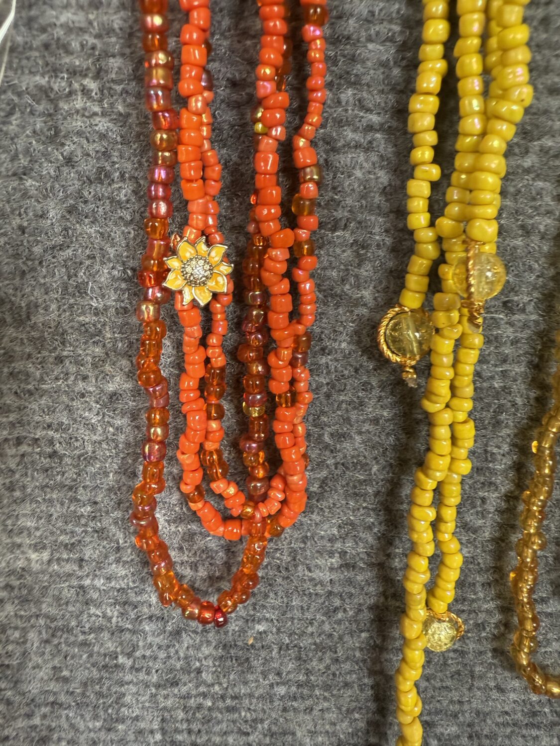Several beaded necklaces in red and orange with a sunflower pendant hanging from one strand, displayed on a gray fabric background.