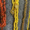 Close-up of colorful beaded necklaces in red, yellow, and amber tones with gold pendants on gray fabric.