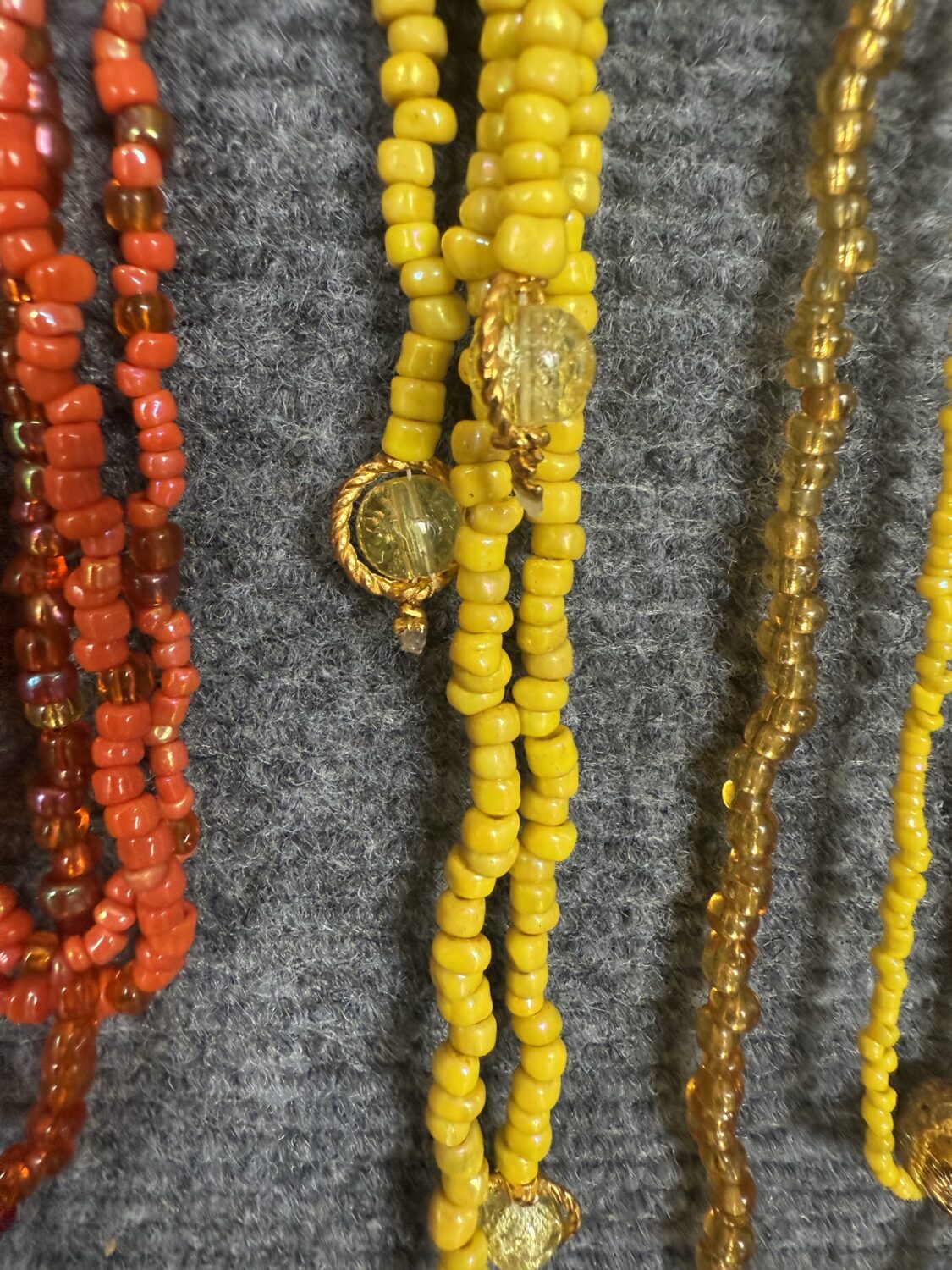 Close-up of colorful beaded necklaces in red, yellow, and amber tones with gold pendants on gray fabric.