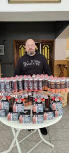 Bearded man with glasses wearing an ALIFE hoodie stands behind a round table filled with rows of labeled cans and boxed gadgets.