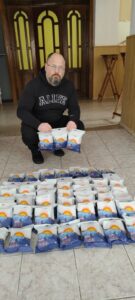 Bearded man in a black ALIFE hoodie kneels beside many blue-and-white packaged goods arranged on the floor, preparing a donation.