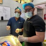 Two surgeons in scrubs, masks, and loupes perform surgery at a draped patient under bright operating lights.