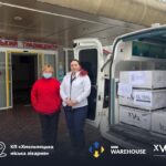 Two women in jackets stand outside a hospital entrance beside an open white van loaded with boxes of medical supplies from humanitarian groups.