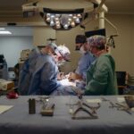 Surgeons in blue gowns and masks performing surgery under bright overhead lights in an operating room, with instruments laid out on a table nearby.