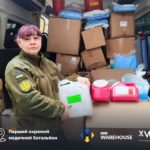 Ukrainian medical battalion member in camo jacket stands beside a van loaded with boxes and supplies, holding a plastic bag.