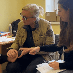 Two women in a small office; one examines an older woman's hand while the other takes notes at a desk cluttered with papers and supplies.