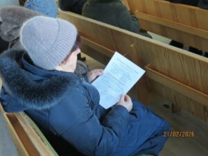 Person in a blue coat with fur hood sits on a wooden bench, reading a printed sheet; date stamp 21/02/2026 visible in corner.