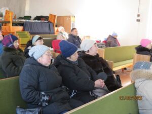 Group of people in winter coats seated on green benches in a bright, simple indoor hall.