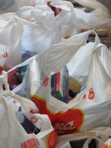 White plastic grocery bags overflowing with groceries, including cereal boxes and cans, on a cluttered surface