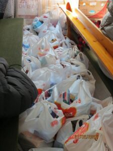 Piles of white plastic grocery bags filled with items stacked along a narrow corridor beside a green sofa and a wooden ramp.