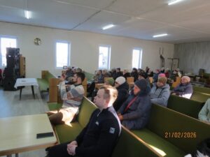 Audience seated in a community hall watching a presentation; a man in the foreground is recording with his phone.