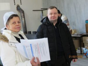 Two people stand in a workshop; a woman in a white coat and headscarf holds papers while a man in a black jacket stands beside her.