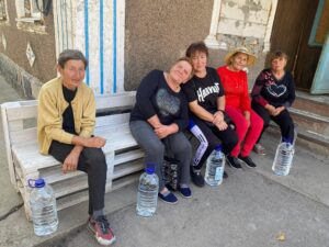 Five women sit on a white wooden bench outside a worn building, with large water bottles at their feet. They wear casual clothing and hats on a sunny day.