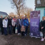 Group of people posing outdoors beside a purple Mobile Care banner; a child stands in front in a rural yard with a leafless tree behind them.