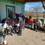 Group of elderly people seated on chairs outside a green-walled house, a man leans over to talk to them in a rural setting.