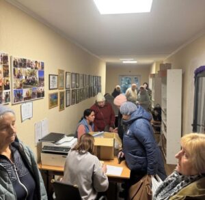 A crowded hallway-style office where people line up at a desk with forms and a staff member assists them, wall displays of photos and certificates in the background.