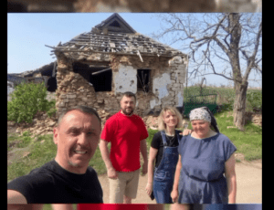 Four people posing for a selfie in front of a crumbling stone house with a broken roof in a rural yard thanks to a toppled wooden frame.