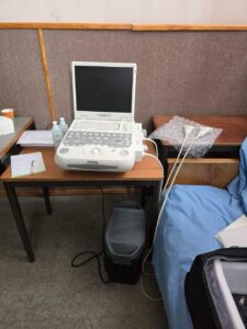 Portable ultrasound machine on a wooden desk in a clinic, with probes on the side and a padded exam bed nearby.