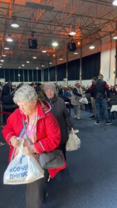 Crowded indoor hall with a metal truss ceiling; people sit and stand in a line holding white plastic bags. In the foreground, an elderly woman in a red jacket walks toward the camera, carrying a bag with Cyrillic text.