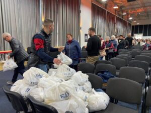 People in a large hall handing out white plastic bags of supplies to a queue of attendees, with stacked bags in the foreground and rows of chairs behind them.