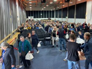 Crowd of people lining up in a large hall to receive bags at a distribution event, many holding white plastic bags and chatting.