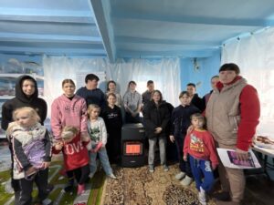 Group of children and adults posing together around a small heater in a blue room with plastic curtains behind them, some wearing jackets and hoodies.