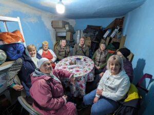 Eight seniors sit around a floral tablecloth in a small, cluttered blue basement room.