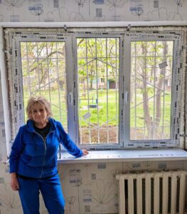 Older woman in a bright blue tracksuit stands by a three-pane window during renovation; outside is a grassy yard with trees and a playground visible, radiator on the right.