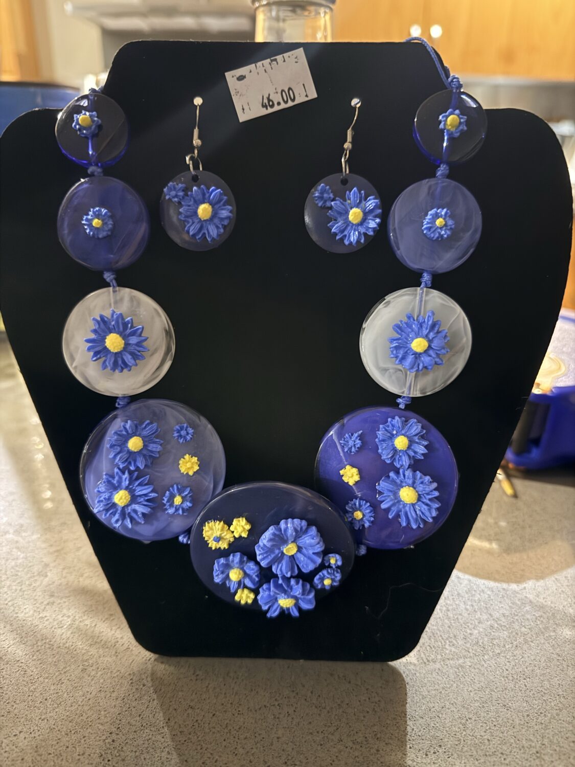 Blue and yellow floral pendant/earring jewelry set on a black display board, featuring round blue discs with blue daisies and yellow centers, connected by cords.