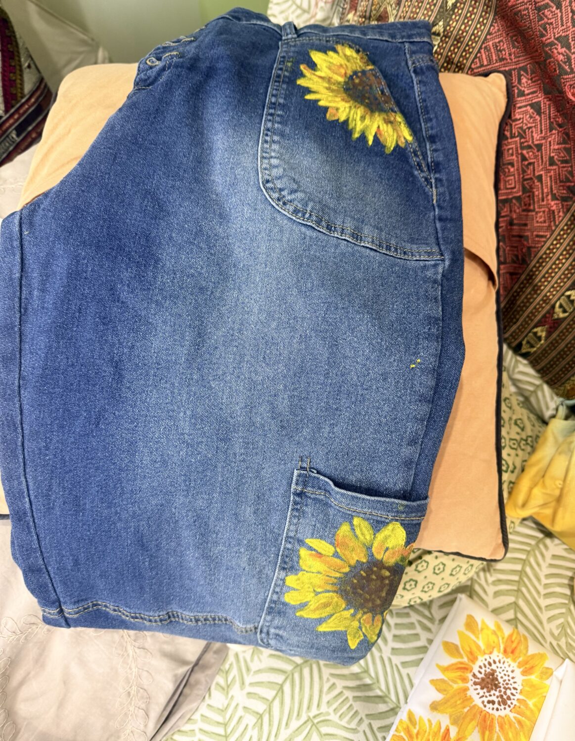 Blue denim jeans folded on a bed with bright sunflower embroidery on the back pocket and upper thigh area.