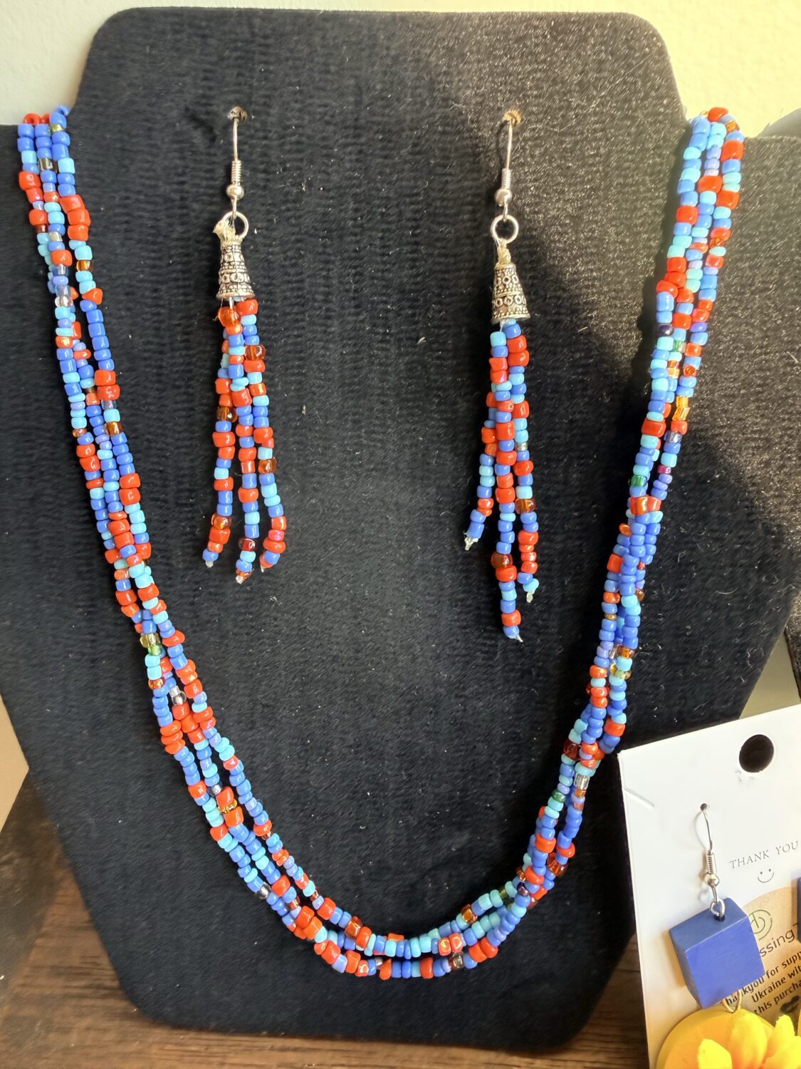 Multi-strand beaded necklace in red, blue, and turquoise with silver bell charms; matching beaded earrings on a black display bust.