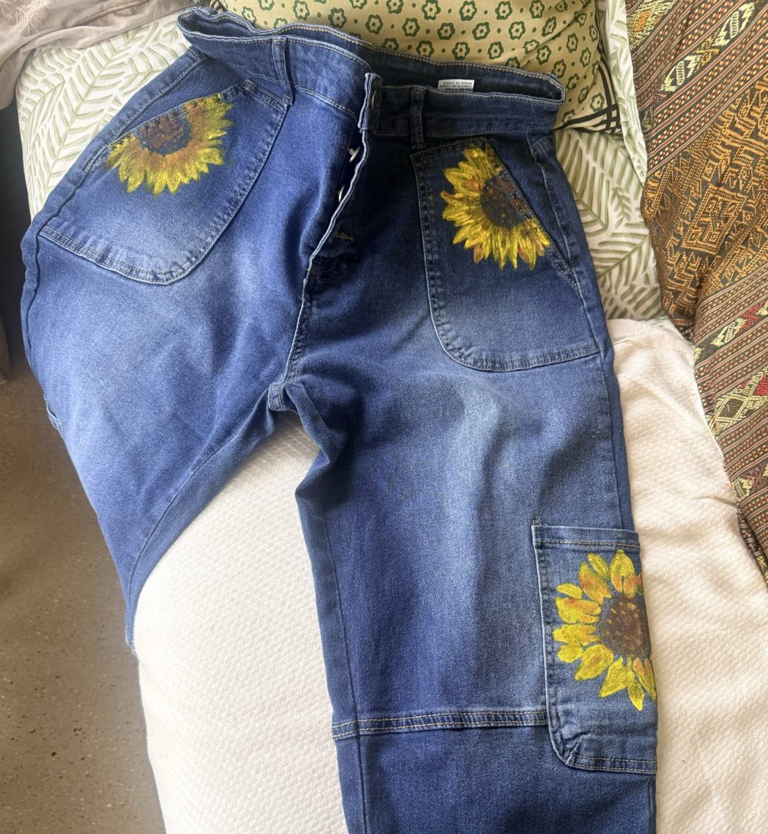 Blue denim jeans with large sunflower embroidery on back pocket and leg pocket detail, laid out on a couch.