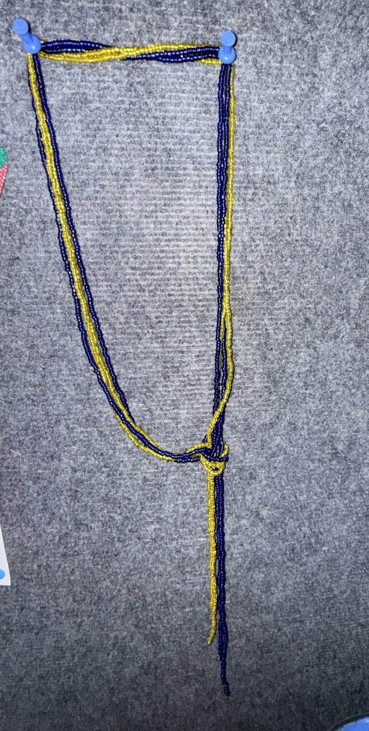 Blue and yellow beaded necklace draped in a U-shape with a knot and hanging strands on a gray fabric background.