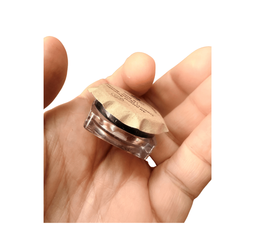 Hand holding a small round cosmetic container with a beige foil seal and a clear base between fingers, against a green background.
