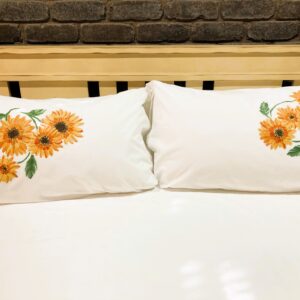 Two white pillows with orange sunflower embroidery on a bed against a wooden headboard and brick wall. Pair of white pillows decorated with orange sunflowers on each side of a bed. Sunflower-embroidered white pillows on a white bedspread. Decorative pillows featuring orange sunflowers on a bed with a brick wall backdrop. Two sunflower-embroidered pillows on a bed.