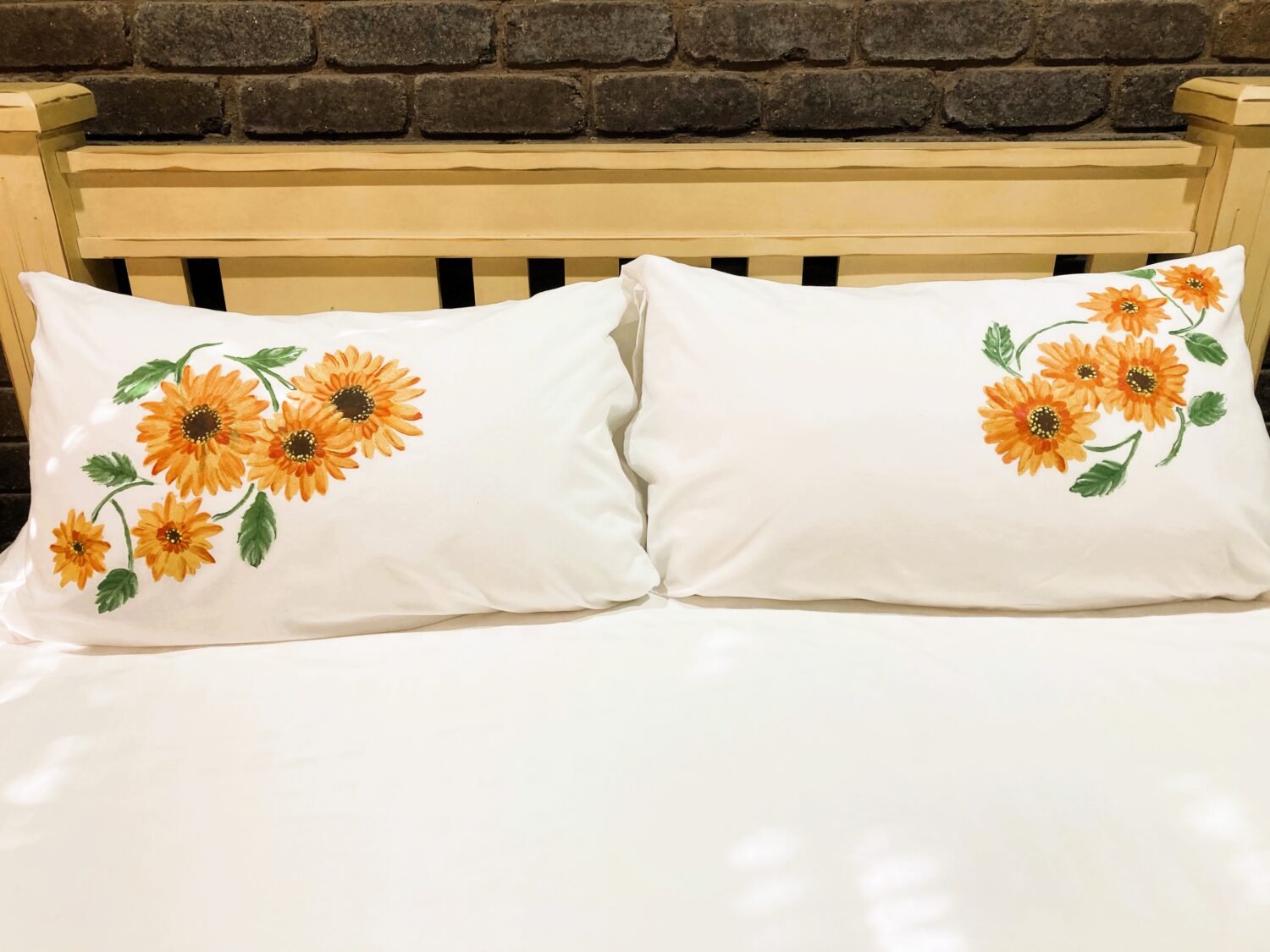 Two white pillows with orange sunflower embroidery on a bed against a wooden headboard and brick wall. Pair of white pillows decorated with orange sunflowers on each side of a bed. Sunflower-embroidered white pillows on a white bedspread. Decorative pillows featuring orange sunflowers on a bed with a brick wall backdrop. Two sunflower-embroidered pillows on a bed.