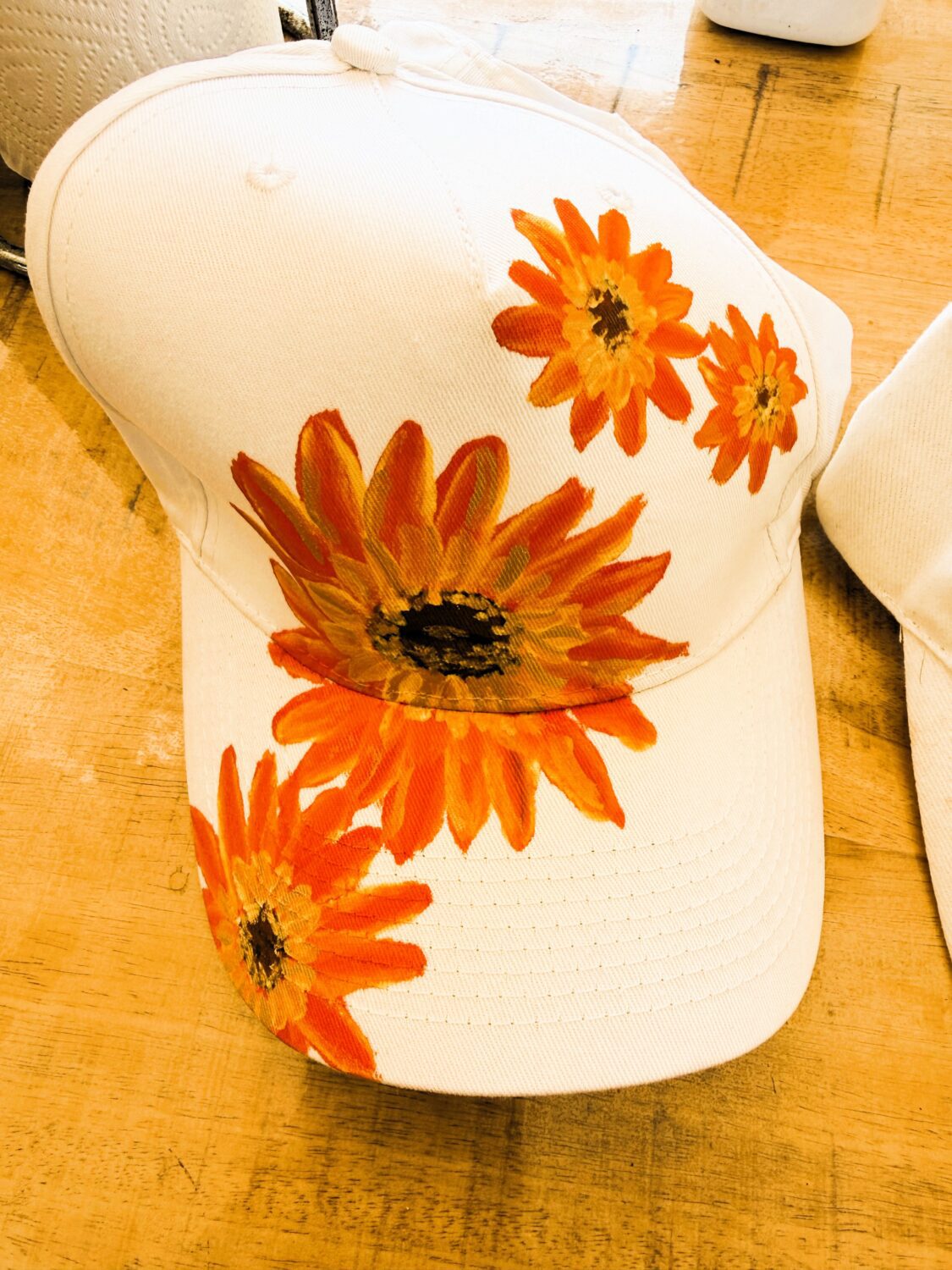 White baseball cap with bright orange sunflower prints on the front and side, resting on a wooden table.