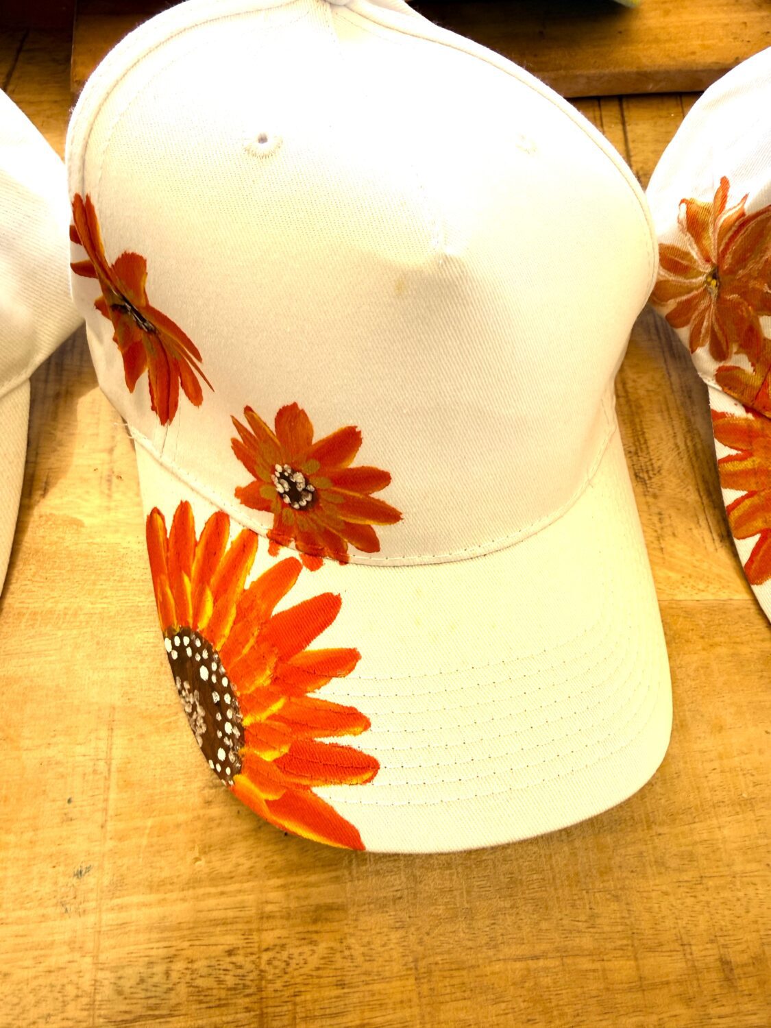 White baseball cap with bright orange sunflower-like flowers painted on the brim and side panels.
