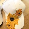 White baseball cap with large yellow sunflowers painted on the brim and crown stitching visible.