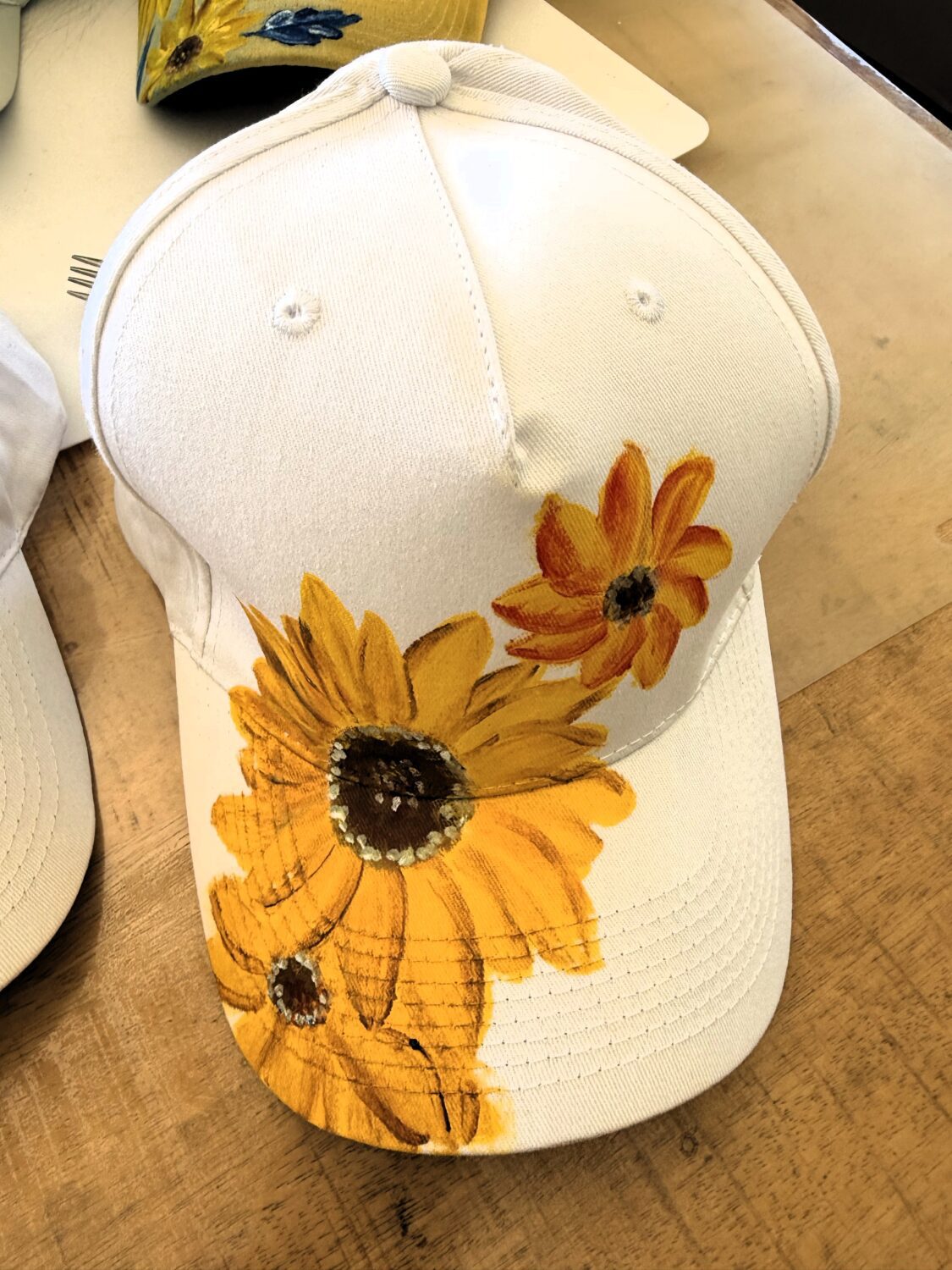 White baseball cap with large yellow sunflowers painted on the brim and crown stitching visible.