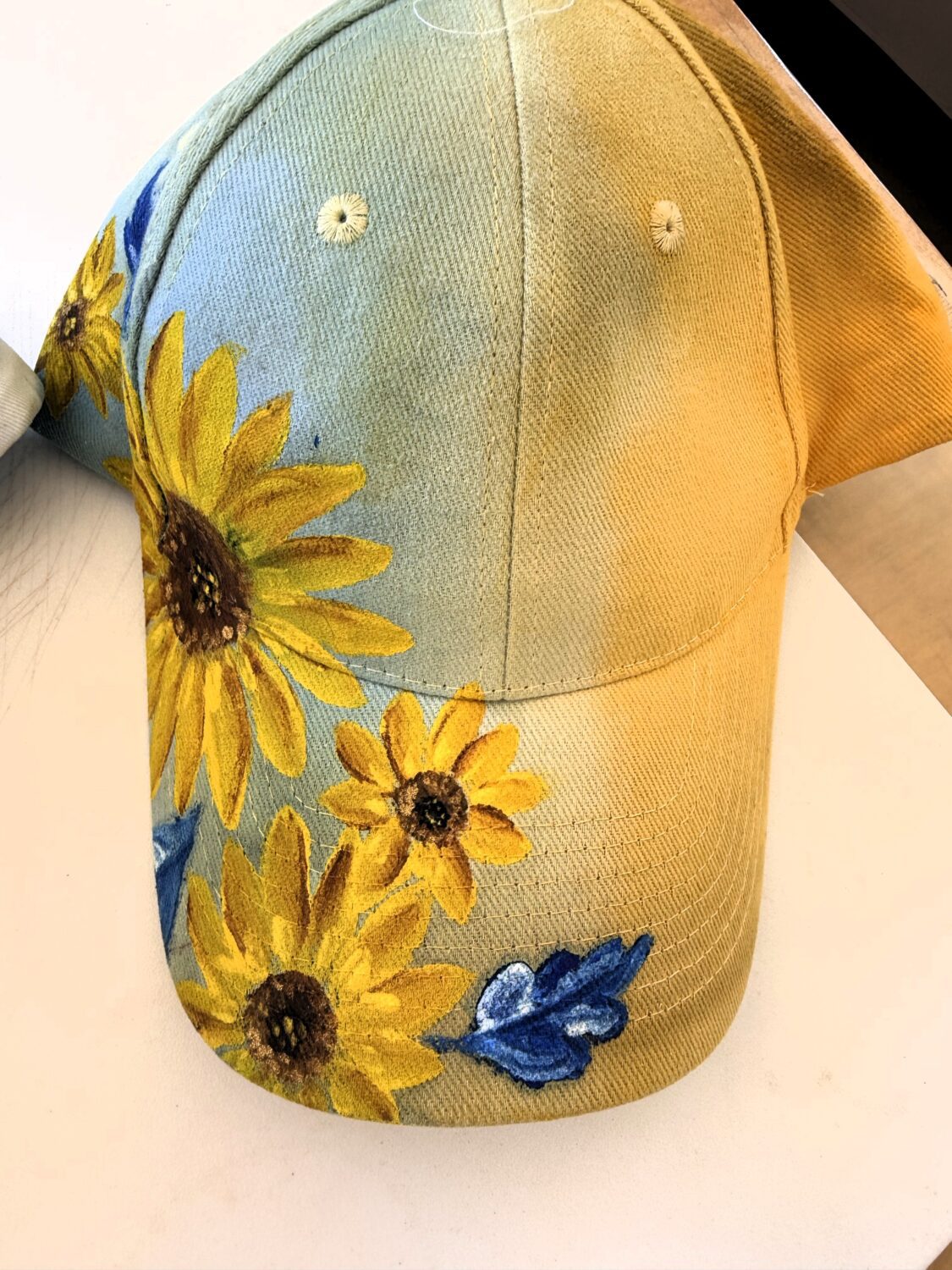 Light blue denim baseball cap with bright yellow sunflower embroidery along the front brim and side panels.