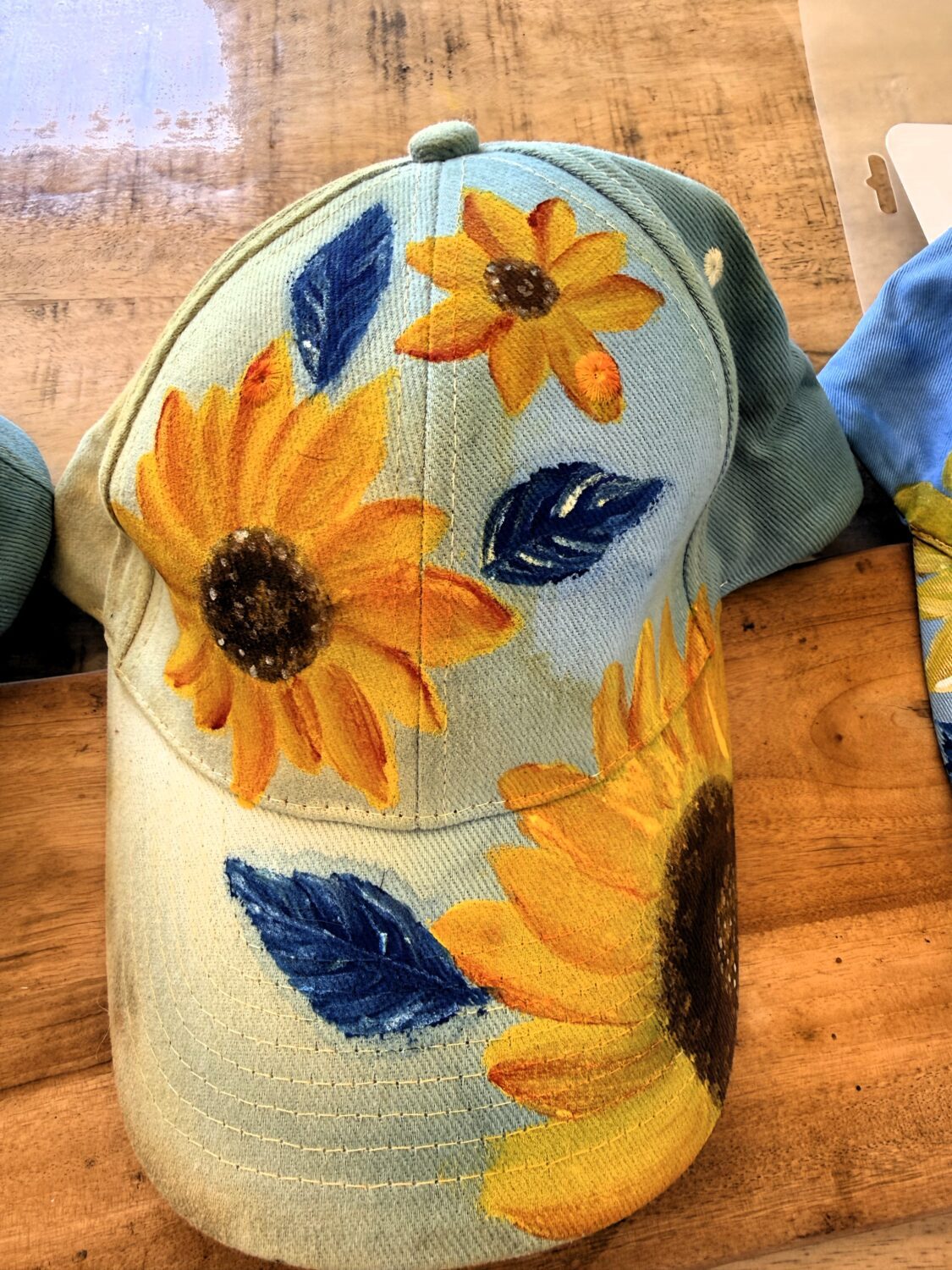 Denim baseball cap with large yellow sunflowers and dark centers on a light blue base, resting on a wooden surface.