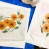 Two white fabric panels printed with bright orange sunflowers and green leaves laid on a blue surface.