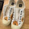 White canvas sneakers with sunflower graphics on the toe caps, placed on a wooden table, no laces. Insole shows VEJA logo.