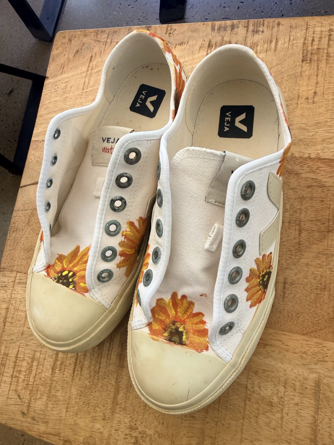 White canvas sneakers with sunflower graphics on the toe caps, placed on a wooden table, no laces. Insole shows VEJA logo.