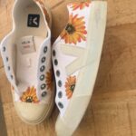 Pair of white canvas sneakers with bright orange sunflower prints, placed on a wooden table, Veja branding visible inside